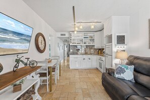 Eine moderne Küche mit weißen Schränken, ein Essbereich mit Holztisch und -stühlen, ein schwarzes Ledersofa und ein großes Gemälde zum Strandthema an der Wand.