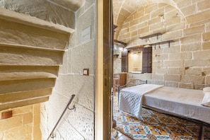 Ein Zimmer mit einem Bett, einer hölzernen Sitzbank, einem Fliesenboden, einer Steinwand und einer Treppe.
