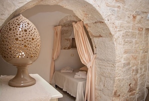 A stone-walled room with a bed, a table with a decorative vase, and a curtain. - Trullo Canicchiulo by BarbarHouse (Latiano)