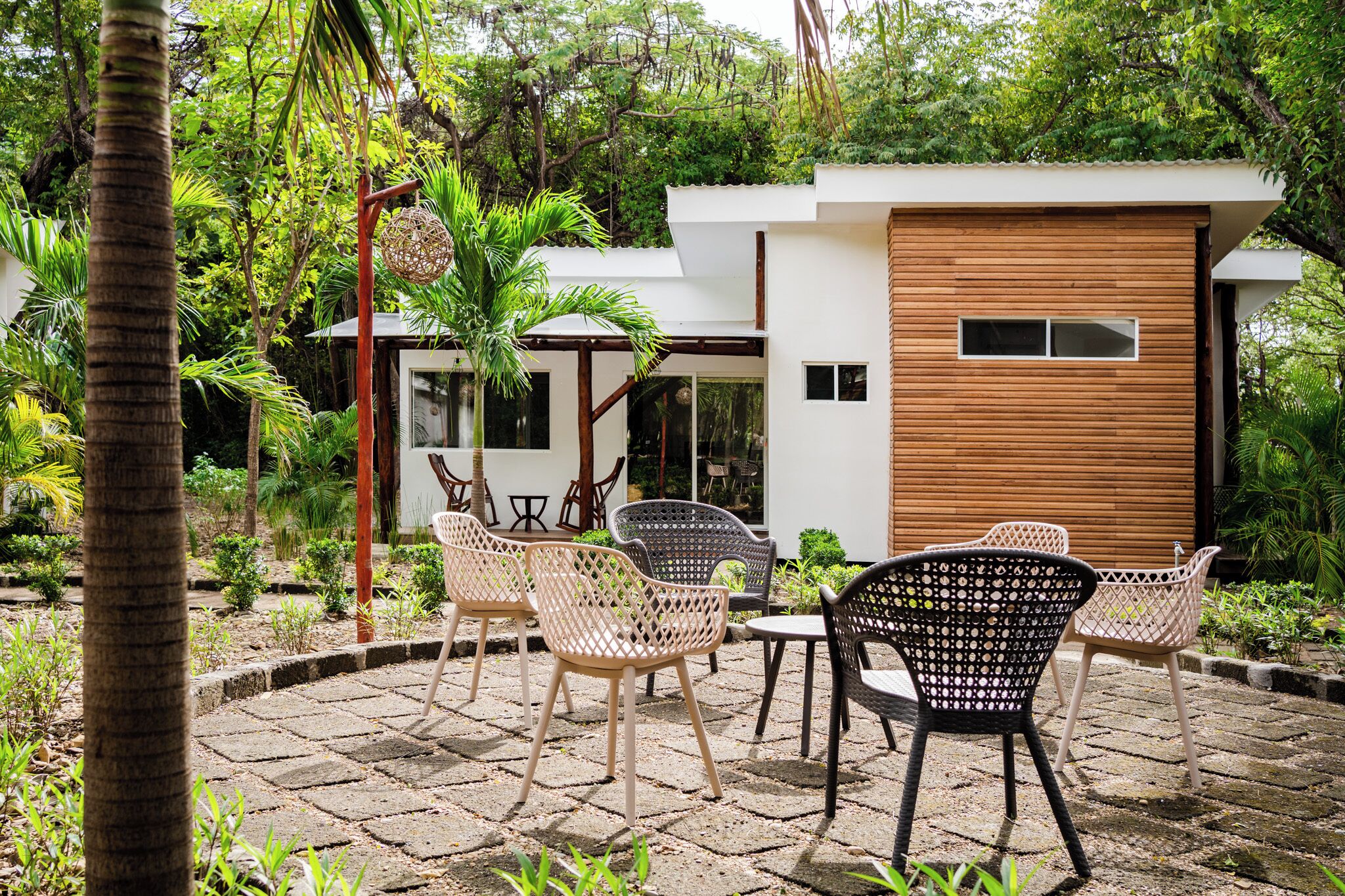 Un espacio moderno al aire libre con sillas de mimbre y una mesa, rodeado de exuberante follaje y un patio empedrado con piedra.