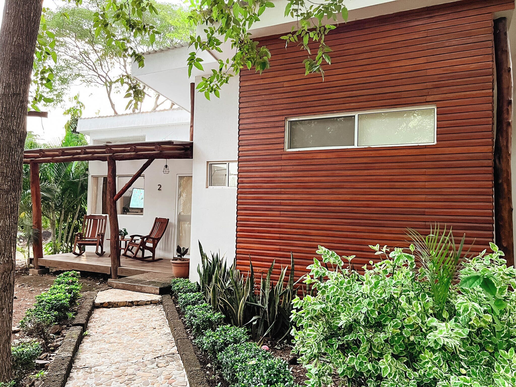 A modern house with a wooden exterior, a covered porch with seating, and a garden with various plants.