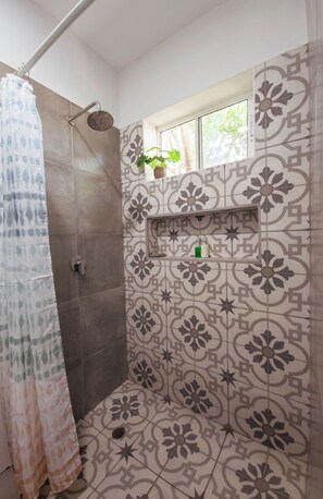 A shower area with patterned tiles, a window with a plant, and a shelf with toiletries. - Ecovillas del Sur (San Juan del Sur)