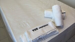 Un lit avec du linge de lit blanc, une serviette pliée sur laquelle on peut lire le texte « VUK APARTMENTS », et un mur sobre avec un interrupteur.