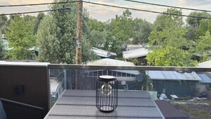Un balcon avec une table et des chaises, un luminaire suspendu, et une vue sur les arbres et les bâtiments.