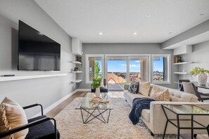 Ein modernes Wohnzimmer mit einer Couch, Couchtisch und Fernseher. Regale mit Dekorationselementen und ein großes Fenster mit Blick auf die Stadt.