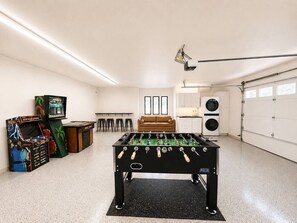 A room with a foosball table, a foosball cabinet, a sofa, a kitchen area, and a garage.