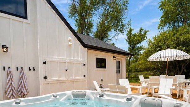 A hot tub in a backyard with a white building, outdoor furniture, and trees. - The Cottage at Tongue River Canyon (Ranchester)