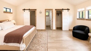 A spacious bedroom with a large bed, a sitting area with a chair, and a view of the outdoors through a window. - The Cottage at Tongue River Canyon (Ranchester)