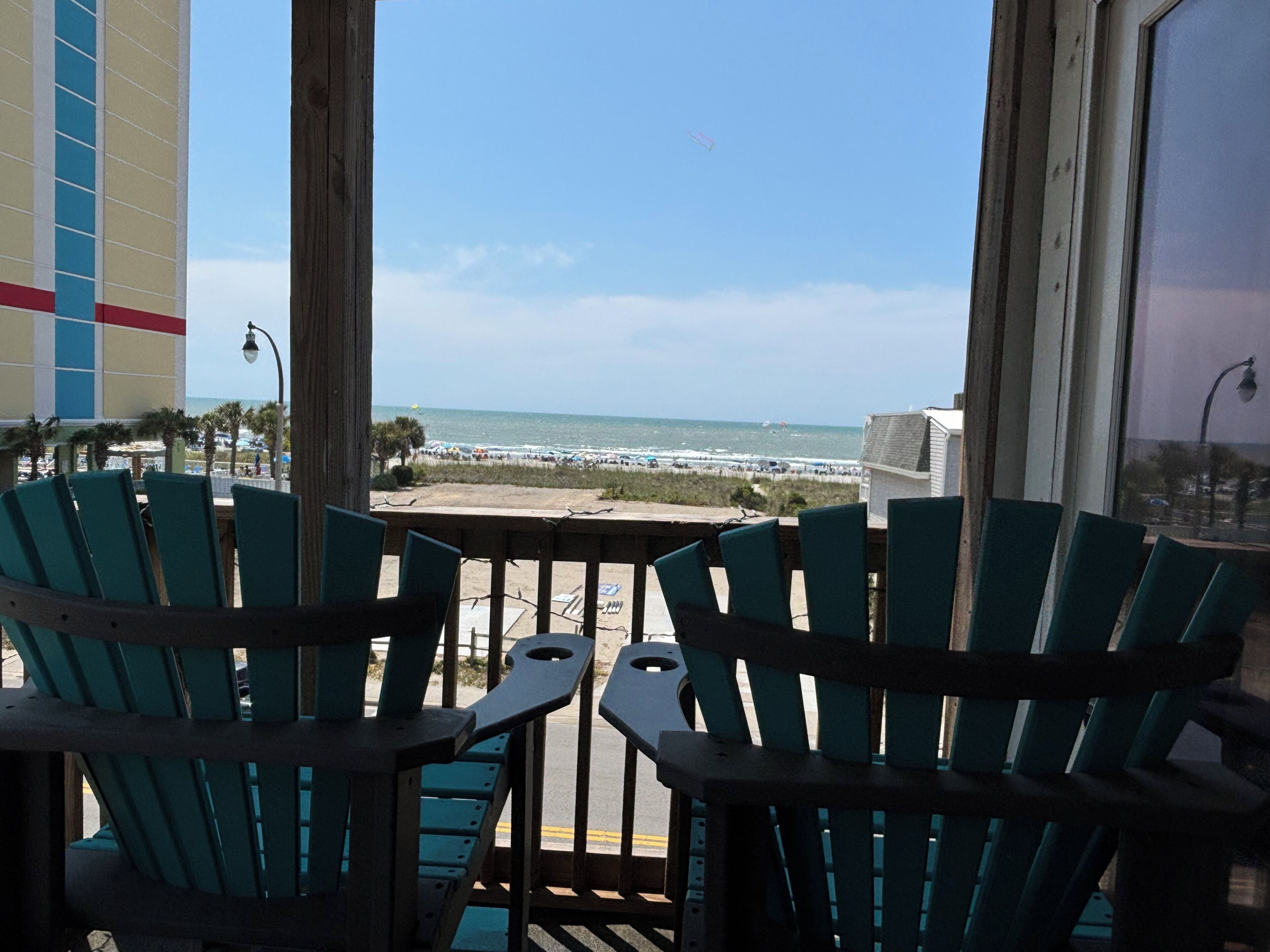 A balcony with green chairs, a view of the beach, and a building with a blue and red facade.