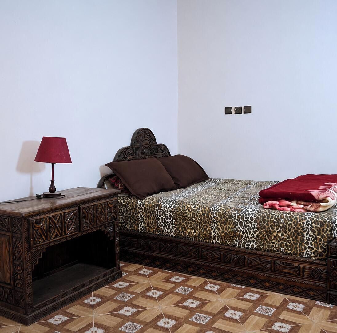 Une chambre à coucher avec une couvre-lit à motifs, une table de chevet en bois surmontée d’une lampe, et un sol à motifs.