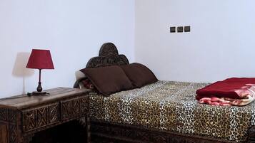Une chambre à coucher avec une couvre-lit à motifs, une table de chevet en bois surmontée d’une lampe, et un sol à motifs.