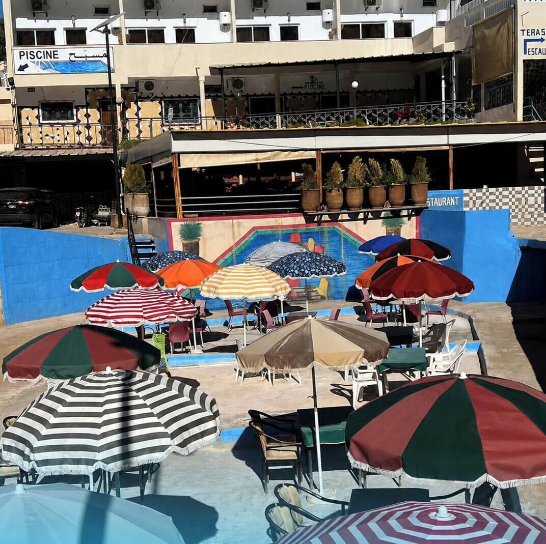 Un área junto a la piscina con múltiples sombrillas y sillas, un restaurante con mesas al aire libre, y un edificio con un cartel que dice 'PISCINE'.