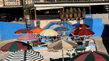 Un área junto a la piscina con múltiples sombrillas y sillas, un restaurante con mesas al aire libre, y un edificio con un cartel que dice 'PISCINE'.