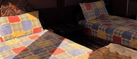 Twee eenpersoonsbedden met geborduurde dekbedovertrekken, een nachtkastje, een lamp en een donker houten hoofdbord.