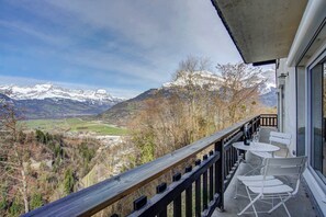 Uma varanda com vista para montanhas cobertas de neve, guarda-corpo de madeira e uma mesa com cadeiras.