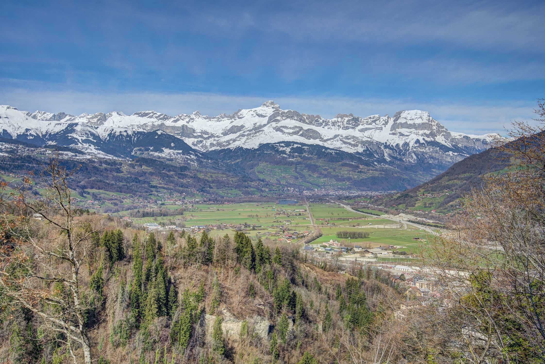 Paisaje montañoso con picos nevados, un valle con edificios y un bosque denso.