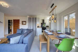 A modern living room with a sofa, dining table, and chairs, a kitchen in the background, and large windows. - Marmottes 3-Bed, 2-Bath - Walk to Spa & Village (Saint-Gervais-les-Bains)