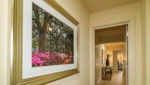 A hallway with a framed picture of a tree-lined path and pink flowers, leading to a room with a door, a table, and a chair. - Suite 33B (Londonderry)