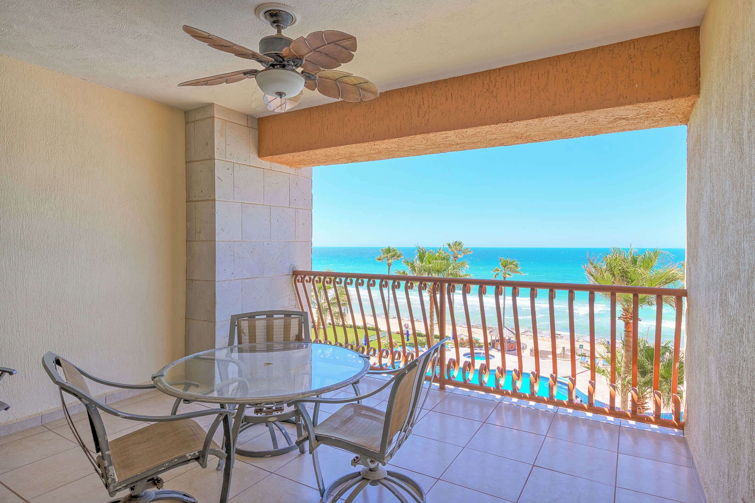 A balcony with a glass table and chairs, a ceiling fan, and a view of the beach.