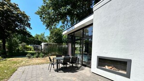 Patio esterno moderno con tavolo e sedie, un elemento decorativo a fuoco e vista su giardino e alberi.