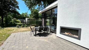 A modern outdoor patio with a table and chairs, a fire feature, and a view of a garden and trees.