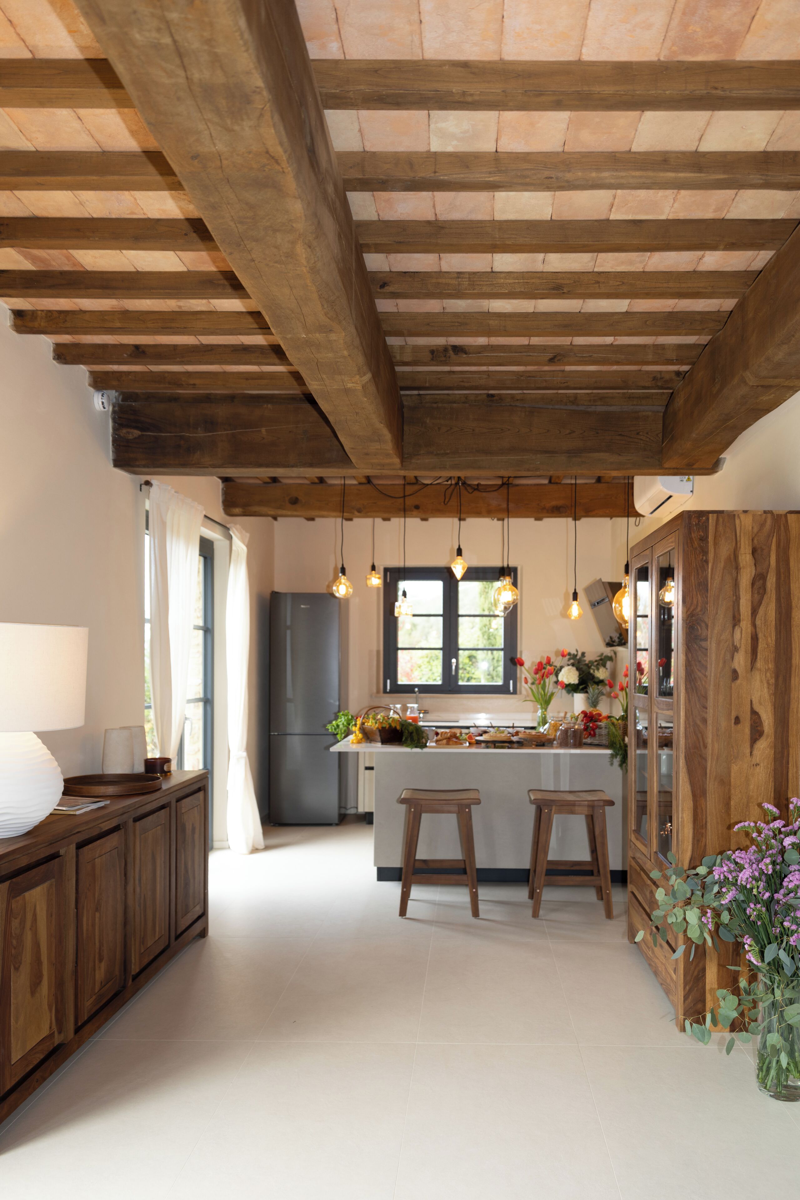 A kitchen with wooden beams, a refrigerator, stools, a window, and hanging lights.