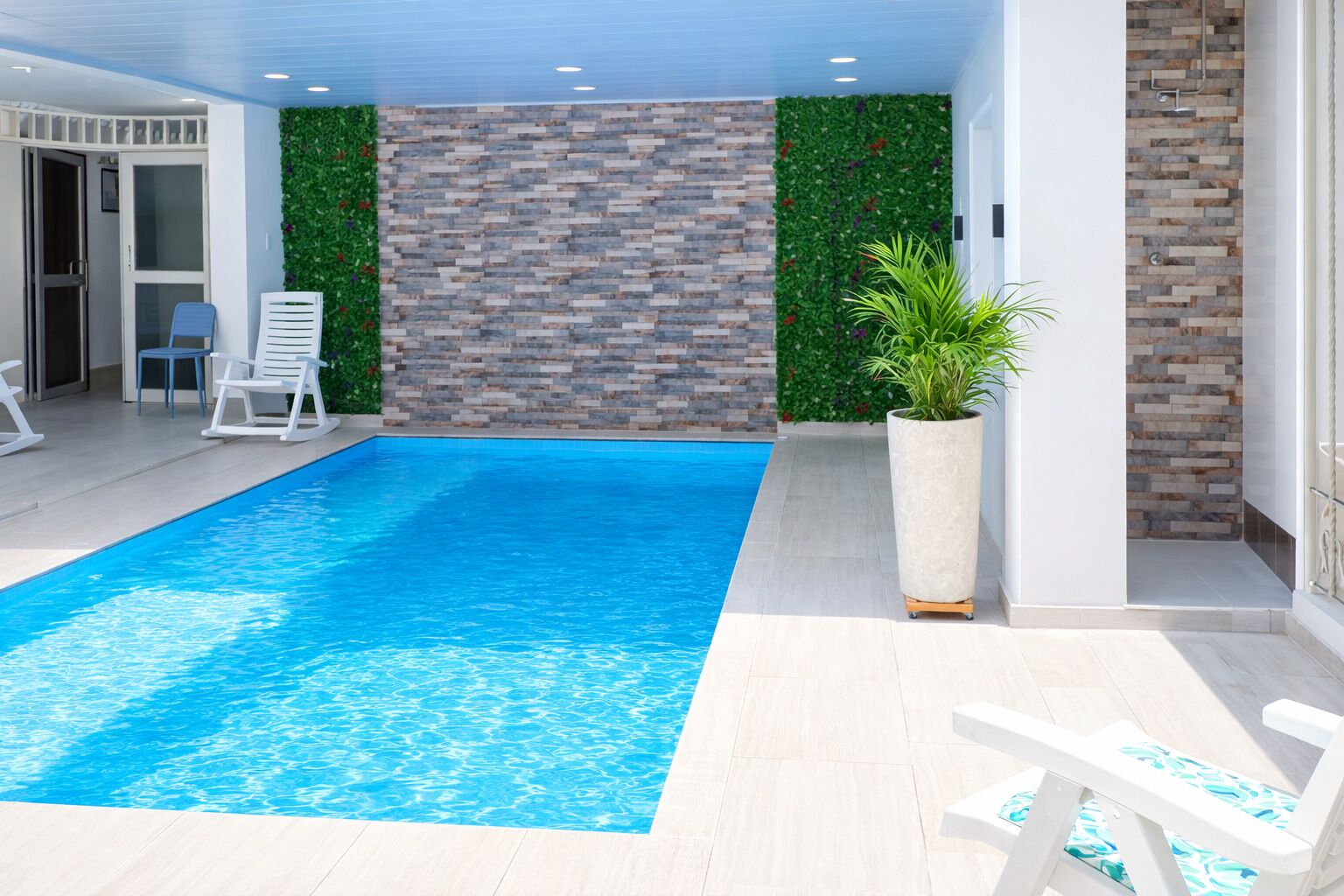 Une piscine avec un sol carrelé, un mur en briques et un mur végétal. On y trouve des chaises longues et un pot de fleurs.