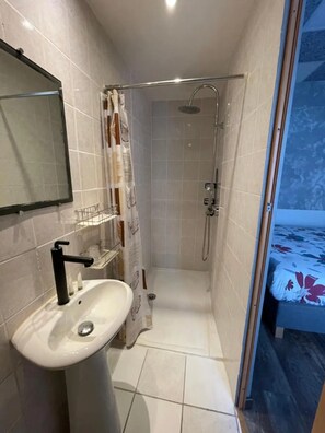 Ein Badezimmer mit Waschbecken, Spiegel, Dusche und einem Bett, das durch eine offene Tür zu sehen ist.