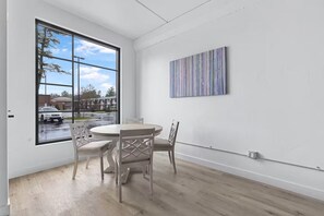 Een kamer met een groot raam, een ronde tafel met stoelen en een abstract schilderij aan de muur.