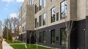 Ein mehrgeschossiges Gebäude mit moderner Architektur, das helle und dunkle Fassadenmaterialien, große Fenster und einen gepflegten Rasen mit jungen Bäumen kombiniert.