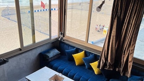 Een kamer met een groot raam met uitzicht op een strand, een blauwe bank met gele kussens, een witte tafel en een vachtkleed.