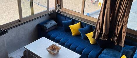 Una habitación con un ventanal grande con vista a una playa, un sofá azul con cojines amarillos, una mesa blanca y una alfombra de piel.