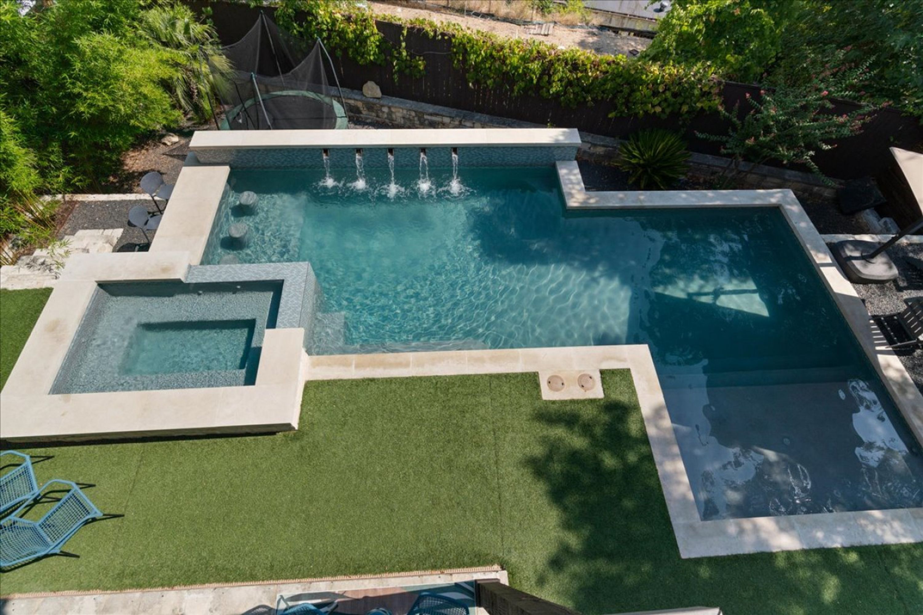 A swimming pool with multiple water features, surrounded by green artificial grass and a stone border.