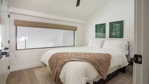 Une chambre avec une grande fenêtre, un lit recouvert d’une couverture marron et deux œuvres d’art vertes accrochées au mur.