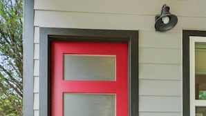 Une porte rouge avec un panneau en verre, un paillasson sur lequel est écrit le mot « MAISON », et une boîte aux lettres noire.