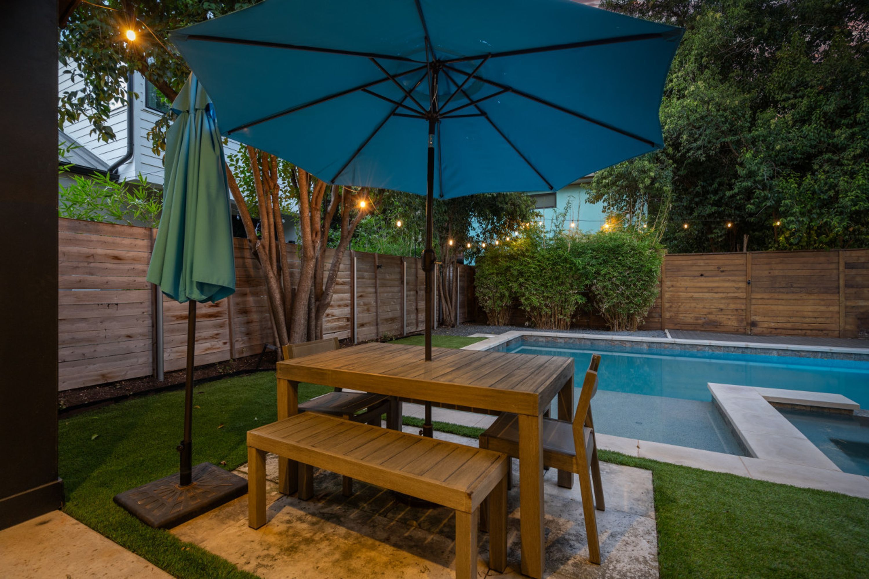 A wooden pool table with an umbrella, a bench, and a pool in a backyard.