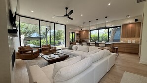 A modern living room with a sectional sofa, a coffee table, and a dining area with wooden cabinets and a large window. - New Build Downtown Views Pool Spa Walkable (Austin)