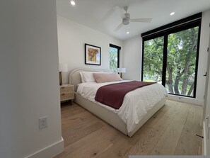 A bedroom with a bed, a ceiling fan, a window with a view of trees, and a bedside table with a lamp. - New Build Downtown Views Pool Spa Walkable (Austin)
