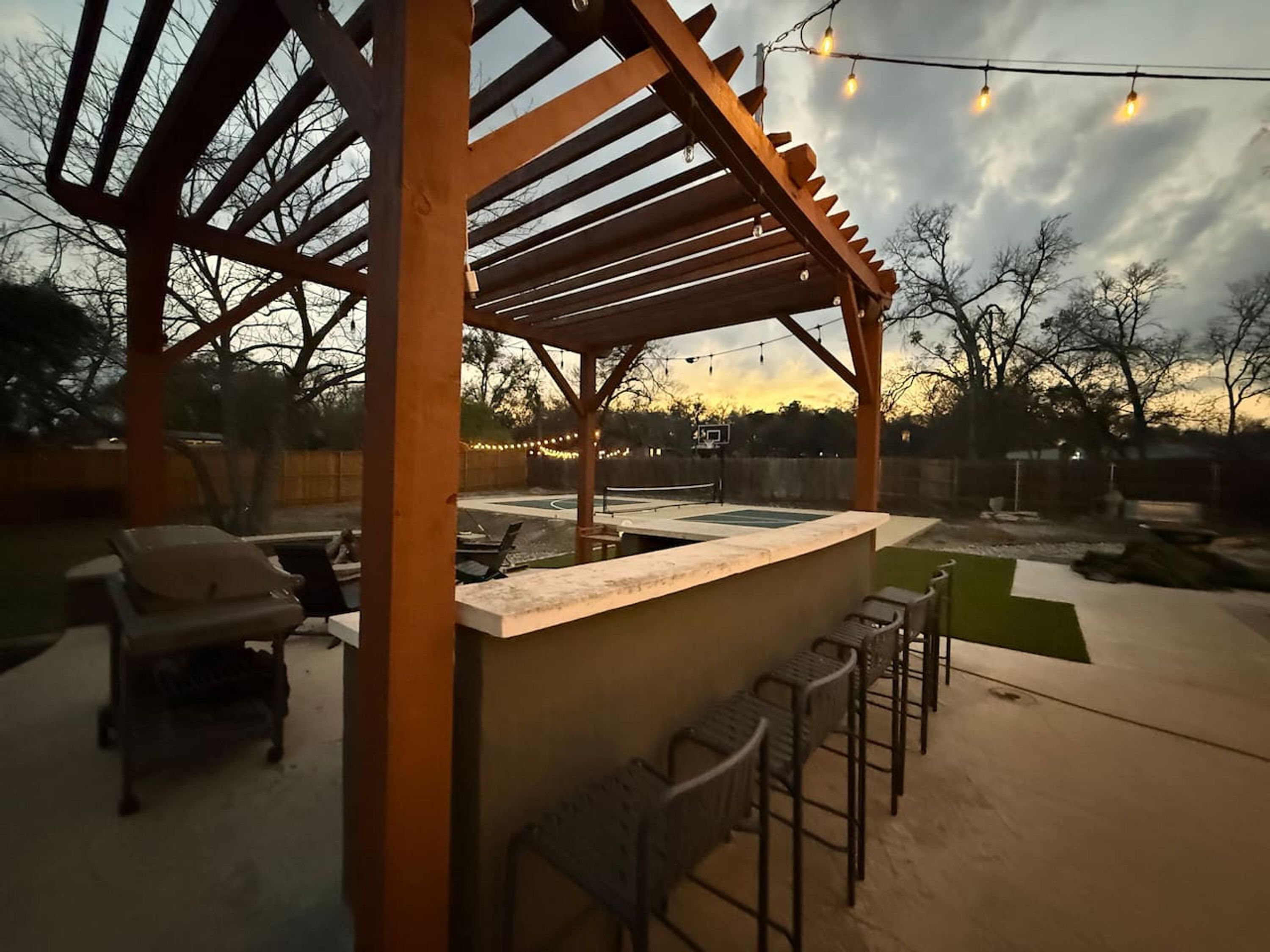 Une terrasse extérieure dotée d’une pergola en bois, d’un coin bar avec des tabourets et d’un panier de basket.