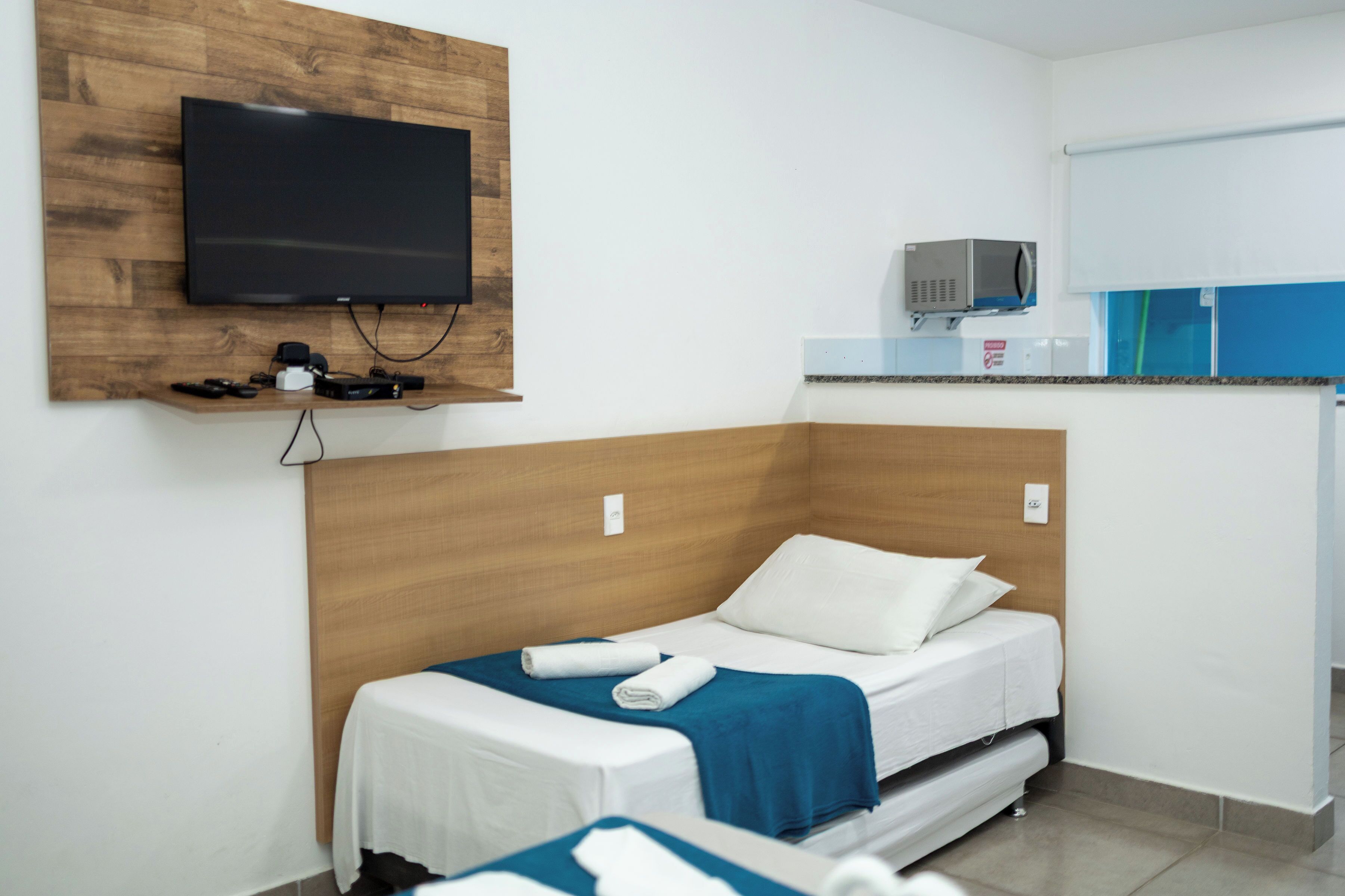 A hotel room with a bed, a television mounted on a wooden panel, a microwave, and a small kitchenette area.