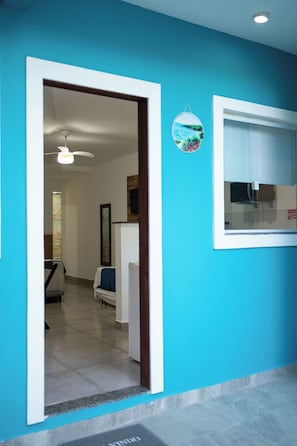 Una habitación con pared azul, una puerta que da a otra estancia, un cuadro enmarco y una ventana con persianas.