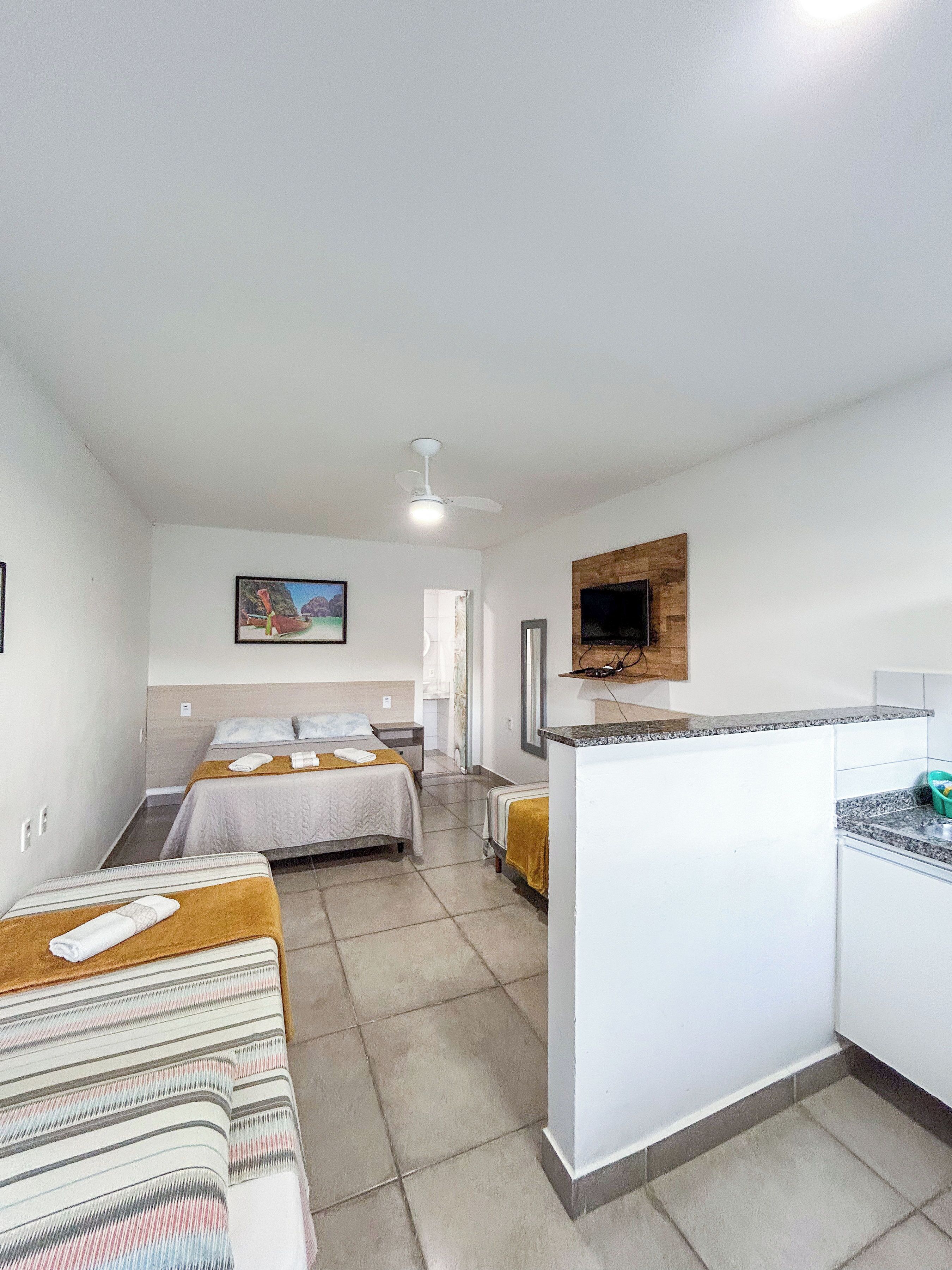 A hotel room with two beds, a kitchenette, a ceiling fan, and a television.
