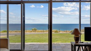 A room with a large sliding glass door leading to a view of the ocean, a lamp on a table, and a television. - South Beach Absolute Beach Front.  Pet friendly! (Carrickalinga)