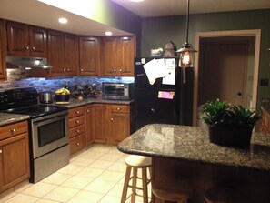Eine Küche mit Holzschränken, einem Edelstahlherd, einer Mikrowelle und einem Kühlschrank. Auf der Arbeitsplatte stehen Weinflaschen und es gibt einen Kübel mit einer Pflanze auf der Kochinsel.