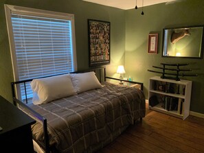 Een slaapkamer met een bed, een raam met zonwering, een wandlamp, een plank met boeken en een opgehangen hertenkop.