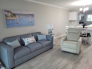 A living room with a grey sofa, a white armchair, a painting of a coastal scene, and a kitchen area with white cabinets and a dining table. - Beachfront Condo Leonardo (Fort Myers Beach)