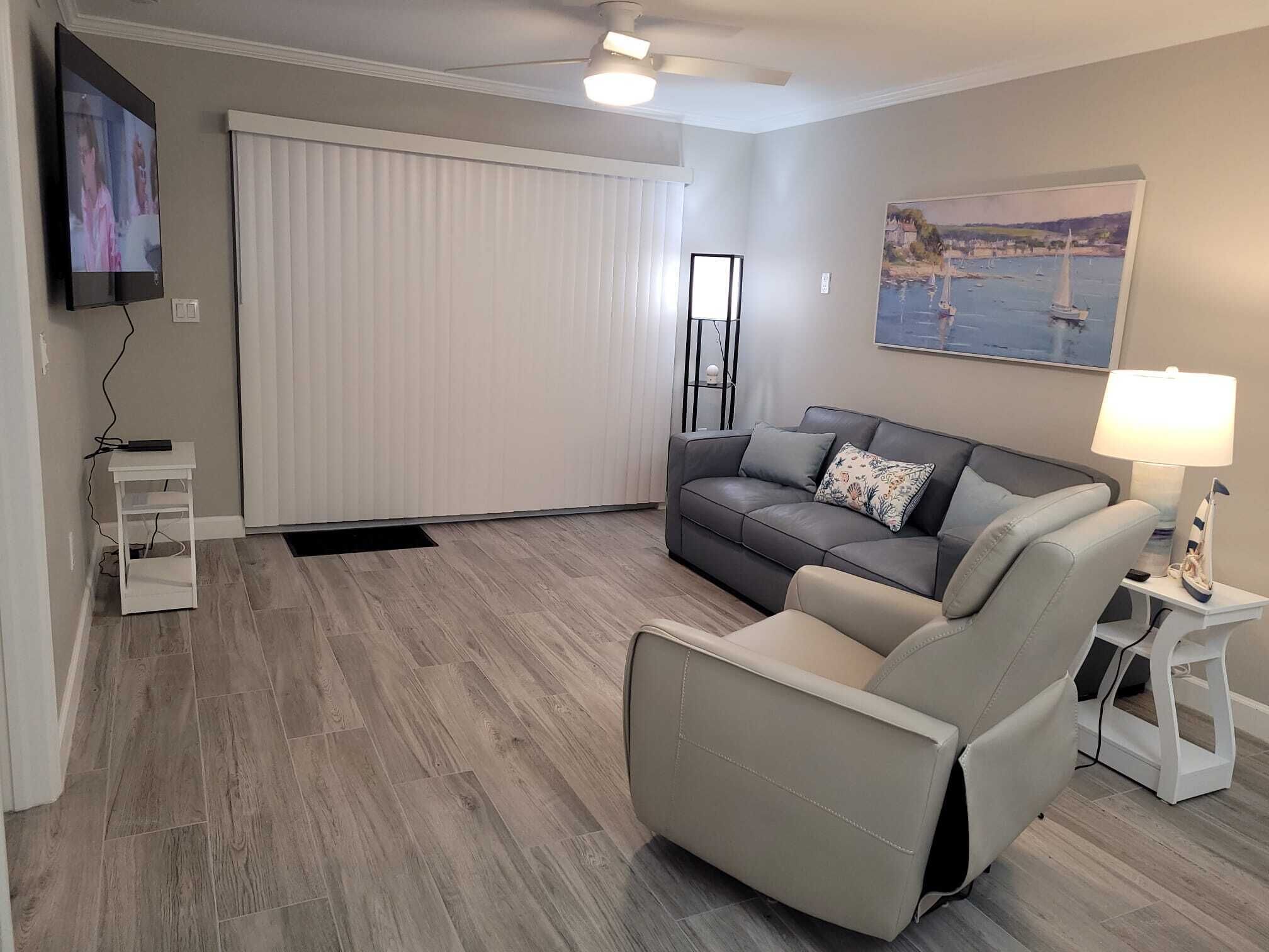 Una sala de estar con un sofá gris, un sillón reclinable, un televisor, un ventilador de techo, un cuadro y una lámpara.