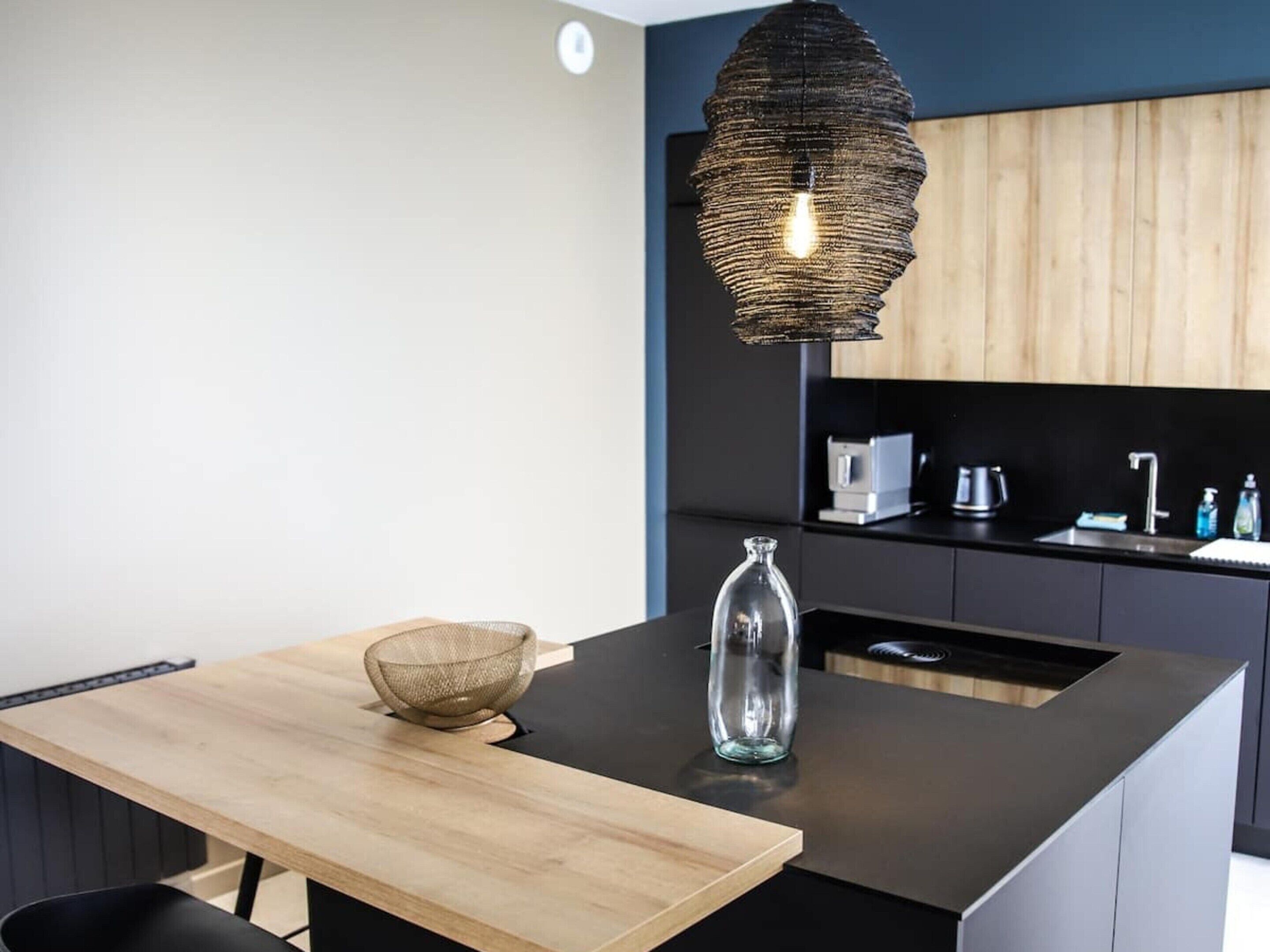 Une cuisine moderne avec un îlot central en bois, des plans de travail noirs, un luminaire suspendu en fibres tressées et un vase en verre.