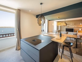 A modern kitchen with a large island, a dining area with a wooden table and bar stools, a TV, and a view of the sea.