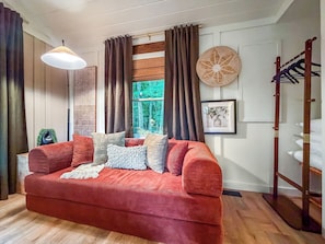 A red sofa with multiple cushions, a hanging lamp, a window with curtains, a woven wall decoration, and a wooden ladder. - Luxe Mtn View 5 Acres, Hot Tub, Screened Porch (Mineral Bluff)